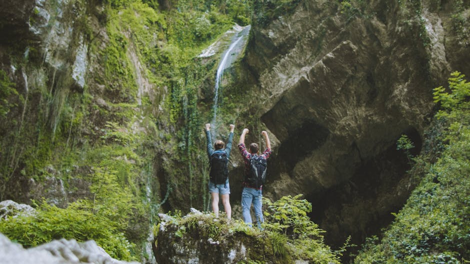 A joyful couple with backpacks raising arms in victory in front of a waterfall amidst lush greenery.